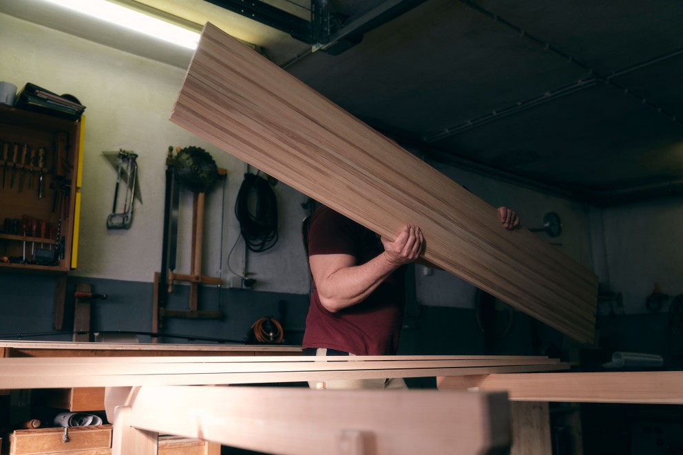 
			Klaus trägt eine lange Holzlatte, den künftigen Skikern, in die Werkstatt.

		