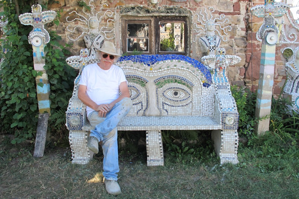 
			Otfried Culmann ruht aus auf der vollendeten „Kopf Bank“, links und rechts der Bank Stelen mit Eulen, ebenfalls aus Mosaik gefertigt

		